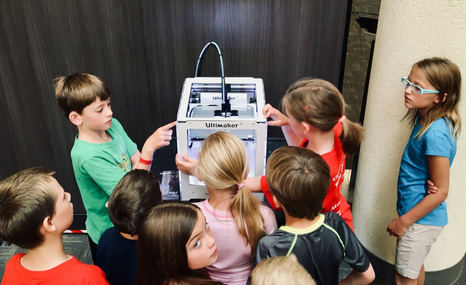 A group of children gather around a 3D printer, watching it work at an after-school STEAM club.
