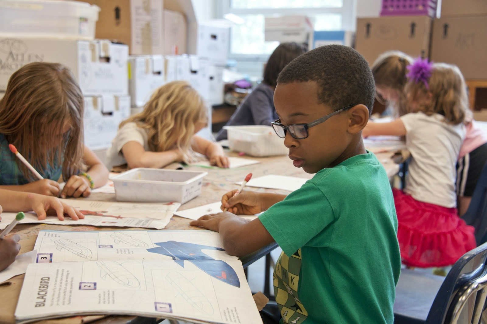 A young boy leans in to study an illustrated book at a busy classroom art table.