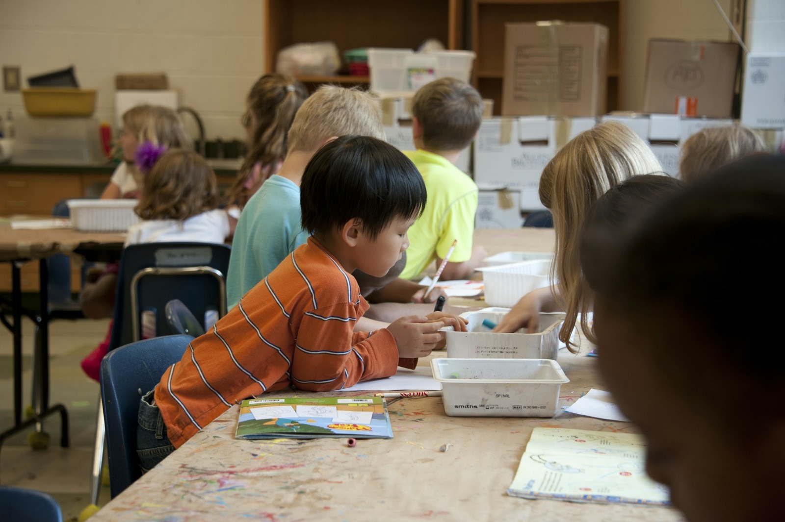 Children work side by side at a classroom art table, drawing and reading together.