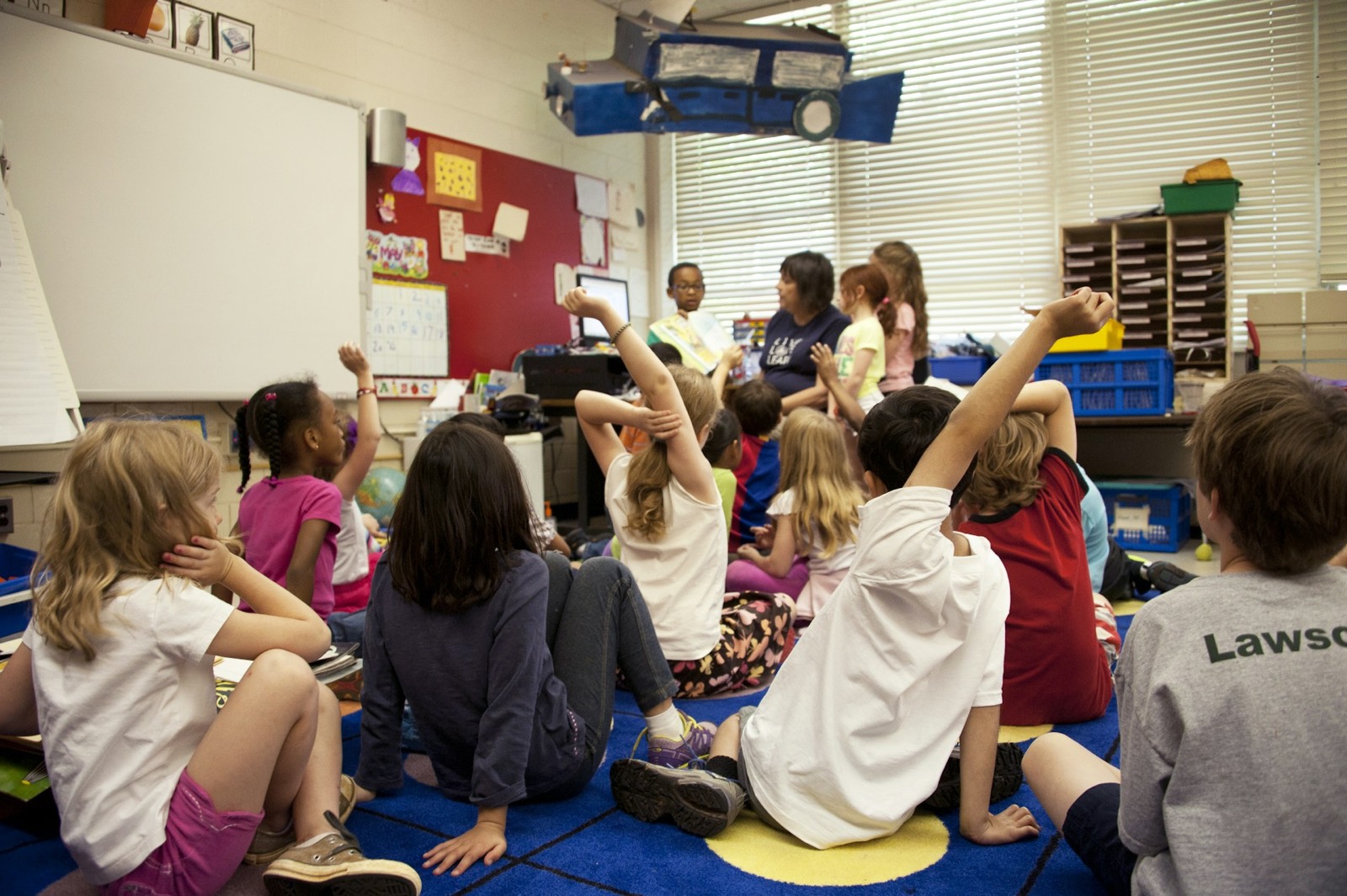 A teacher reads a picture book aloud to a circle of children who eagerly raise their hands.